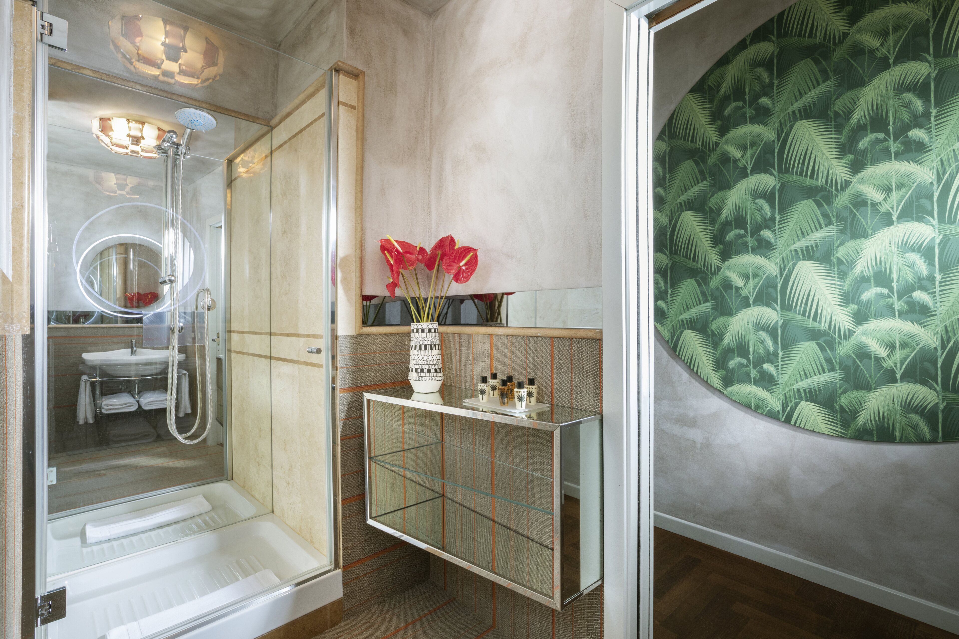 A bathroom with a glass shower enclosure, a sink, a vanity with a mirror, and a decorative wall with a palm tree pattern.