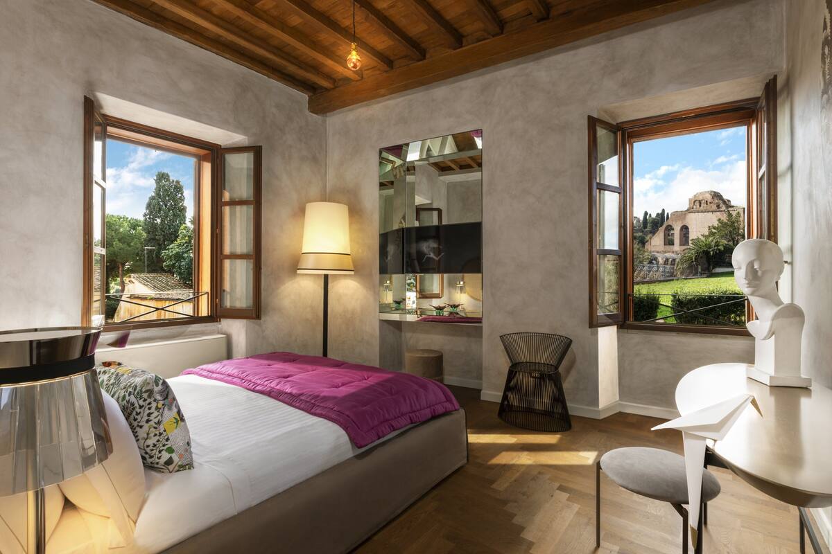 Une chambre à coucher comprenant un lit, une lampe de chevet, une chaise, une table et une vue sur un jardin et un bâtiment, le tout visible à travers de grandes fenêtres.