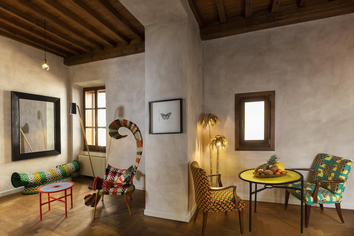 Une pièce avec un plafond en bois, une table ronde avec un plateau jaune, deux chaises à motifs, un tabouret rouge et un tableau encadré accroché au mur.