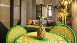Een eetgedeelte met een ananas als middelpunt, groene geweven stoelen, een groene bank met patroon kussens en een hoge vloerlamp.