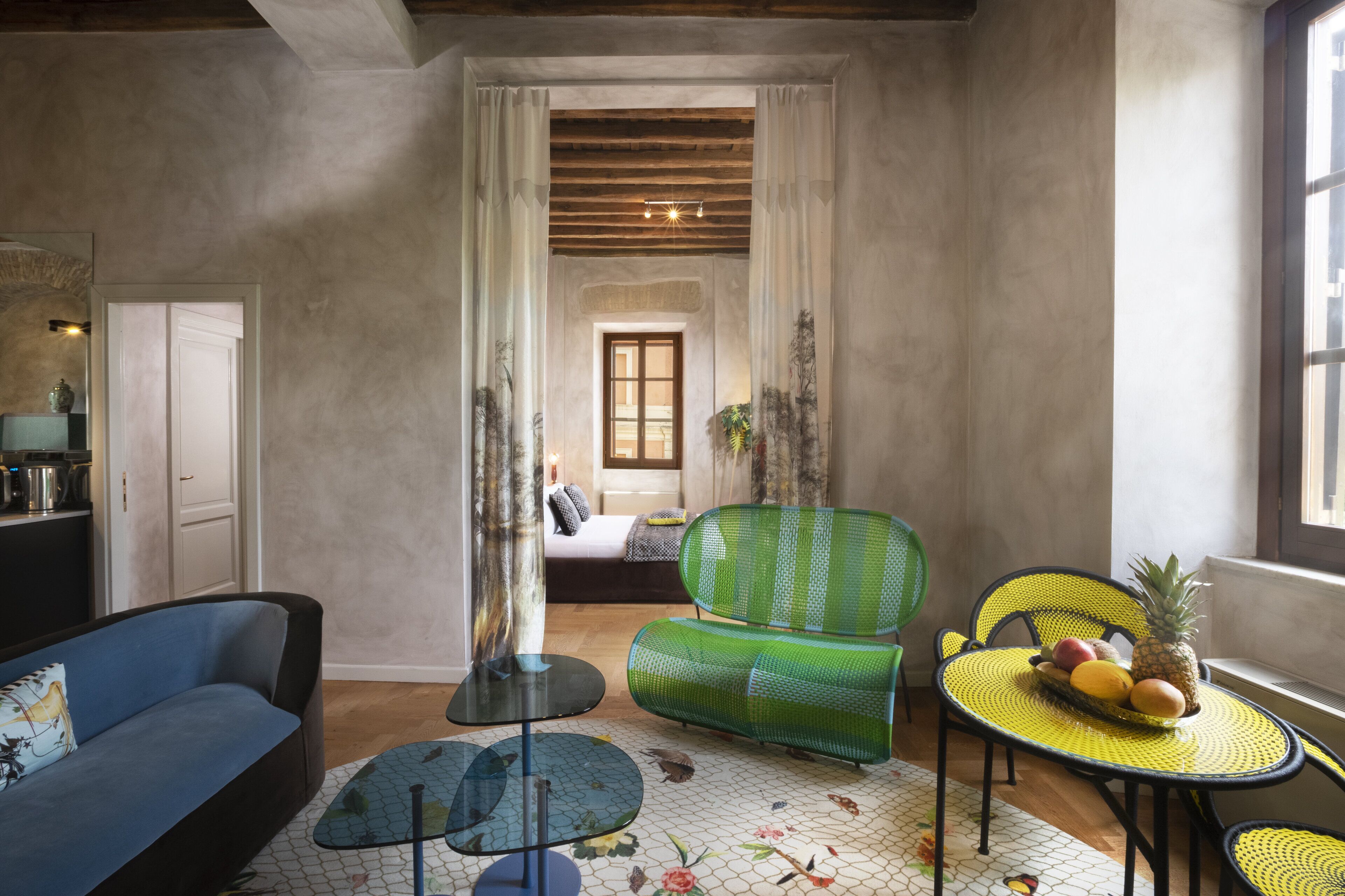 Una sala moderna con sofá, sillón verde y una mesa amarilla con fruta. Se ve una cama y una ventana al fondo.