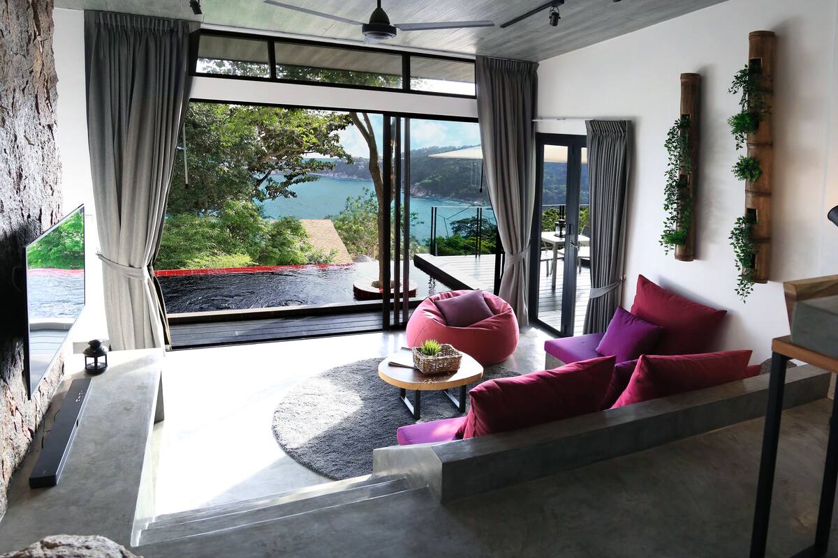 Ein modernes Wohnzimmer mit einem großen Fenster, das Blick auf ein Gewässer und Grünflächen bietet. Es gibt eine Sitzecke mit einem runden Tisch und Kissen.