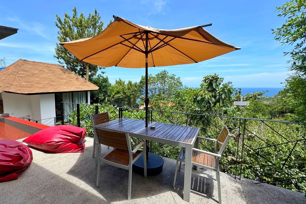 Eine Terrasse mit Tisch und Stühlen, einem großen Sonnenschirm und Blick auf den Ozean.
