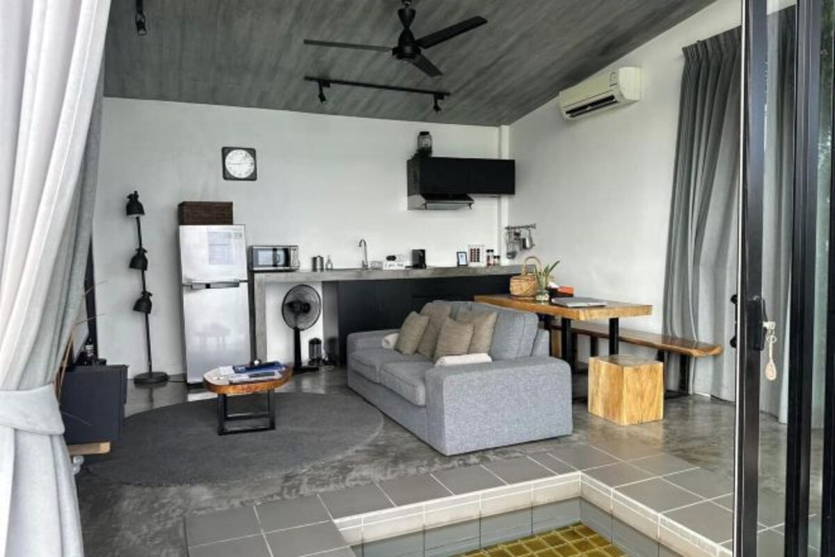 Una sala moderna con un sofá gris, una mesita de madera, un ventilador de techo y una zona de cocina con microondas, fregadero y armarios.
