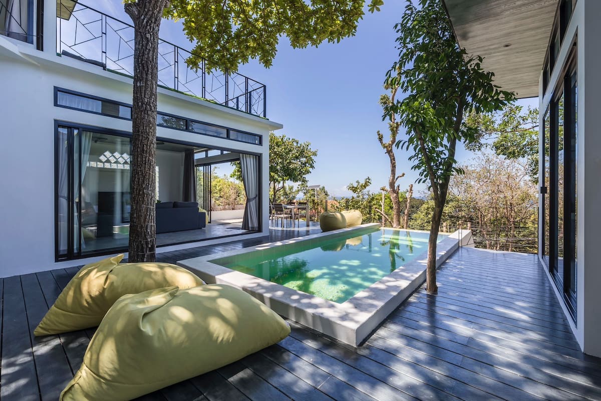 Ein moderner Poolbereich im Freien mit einer Holzterrasse, einer integrierten Sitzgruppe und einer gläsernen Struktur.