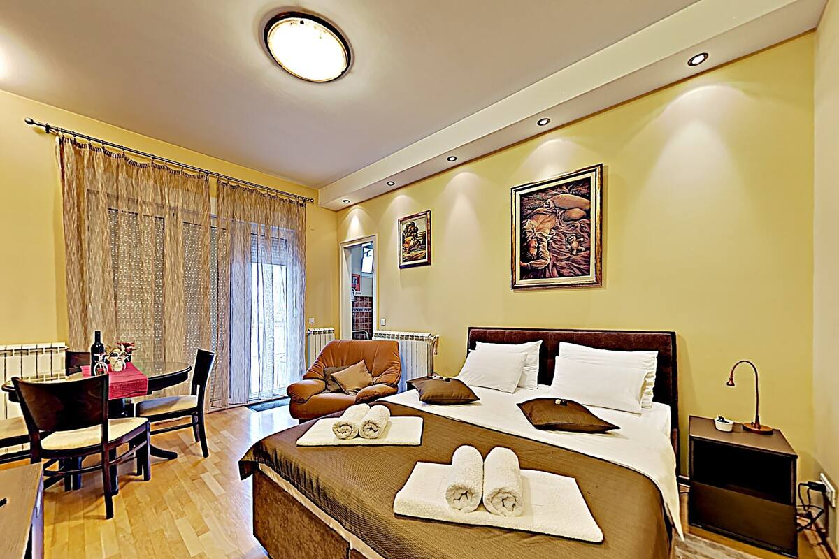 A hotel room with a large bed, a sitting area with a table and chairs, a window with curtains, and artwork on the walls.