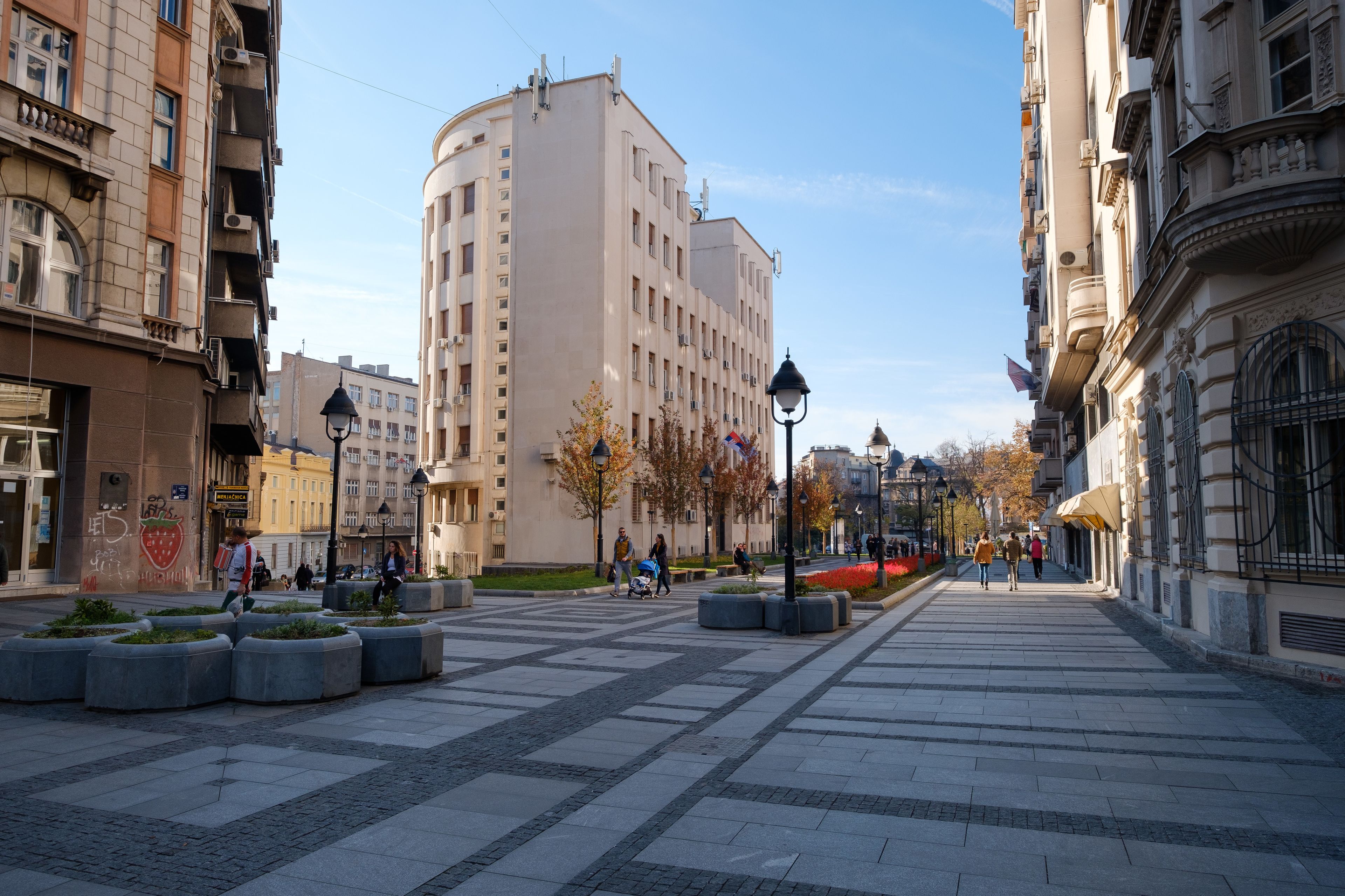 Eine breite Fußgängerzone mit modernen Gebäuden, einem zentralen Platz mit Pflanzkübeln und blauem Himmel.