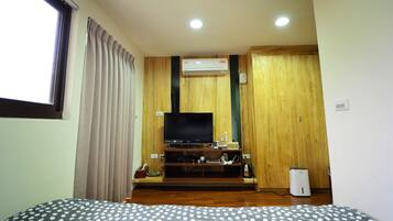 A hotel room with a wooden floor, a bed with a polka dot bedspread, a television on a wooden stand, an air conditioning unit, and a window with curtains.