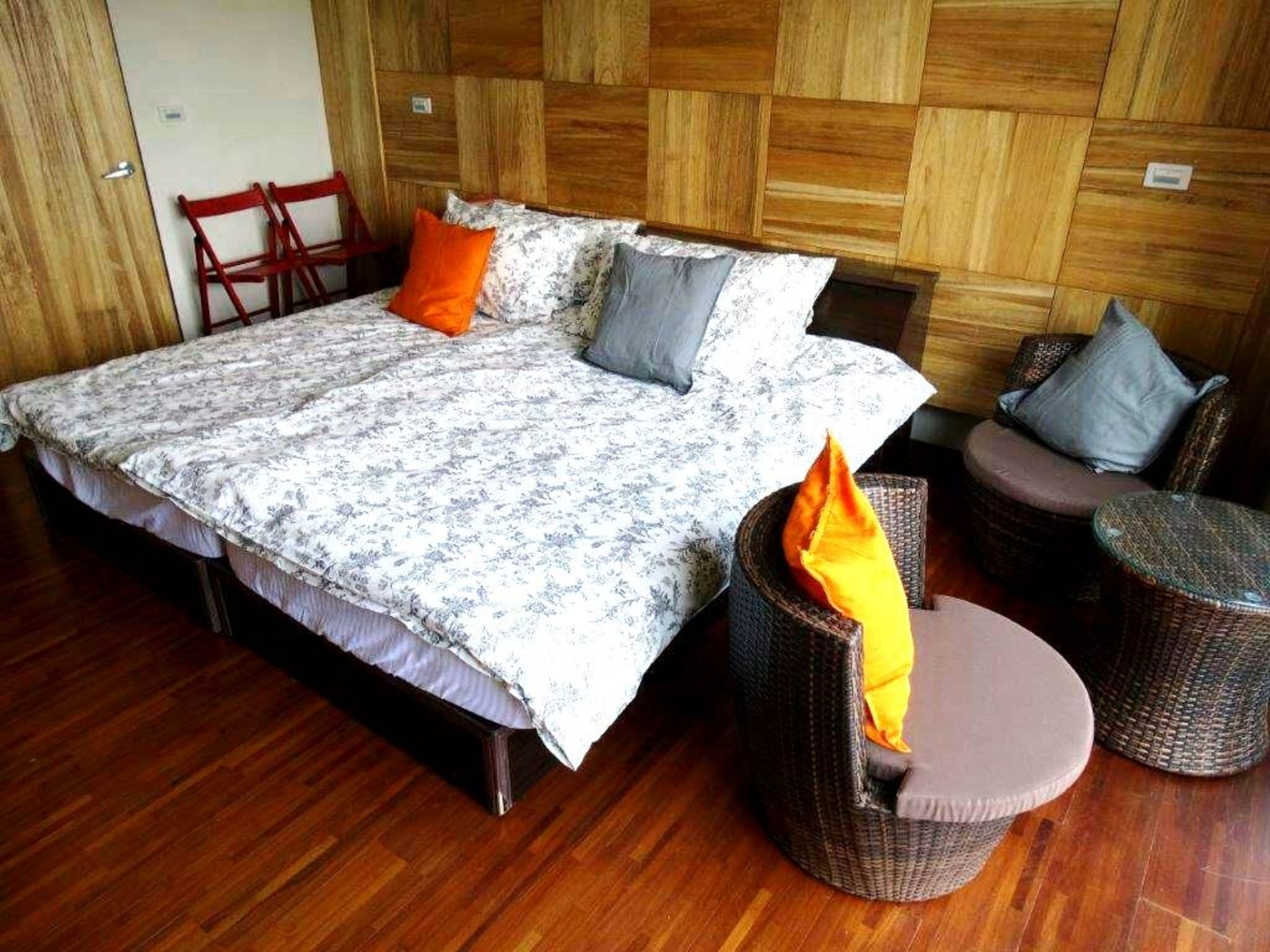 Een groot bed met een bloemenbedekking, twee kussens en een nachtkastje. Er staan rieten stoelen bij en een klein tafeltje met kussens.