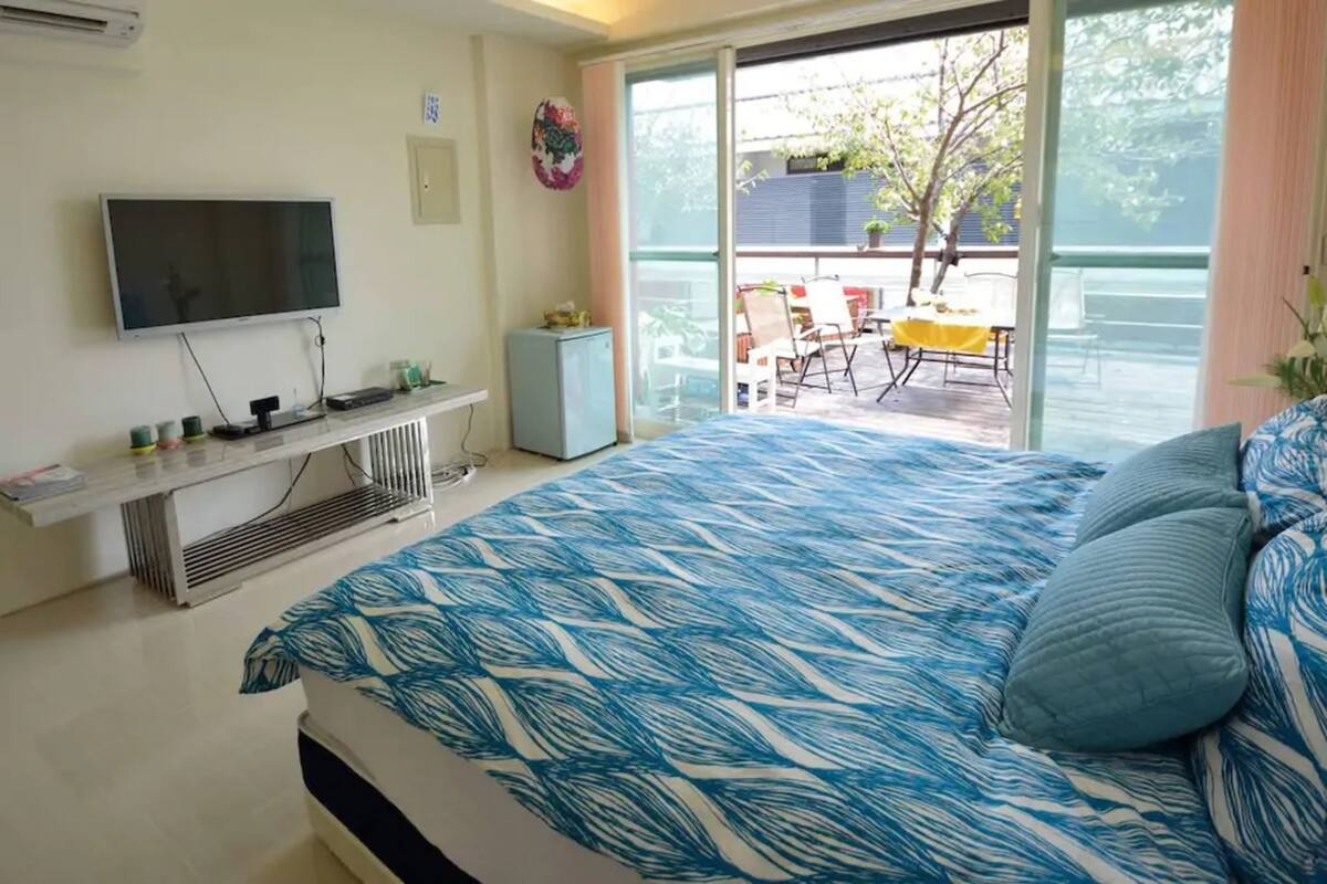 Quarto com cama, televisão, um pequeno armário e varanda com cadeiras e mesa.