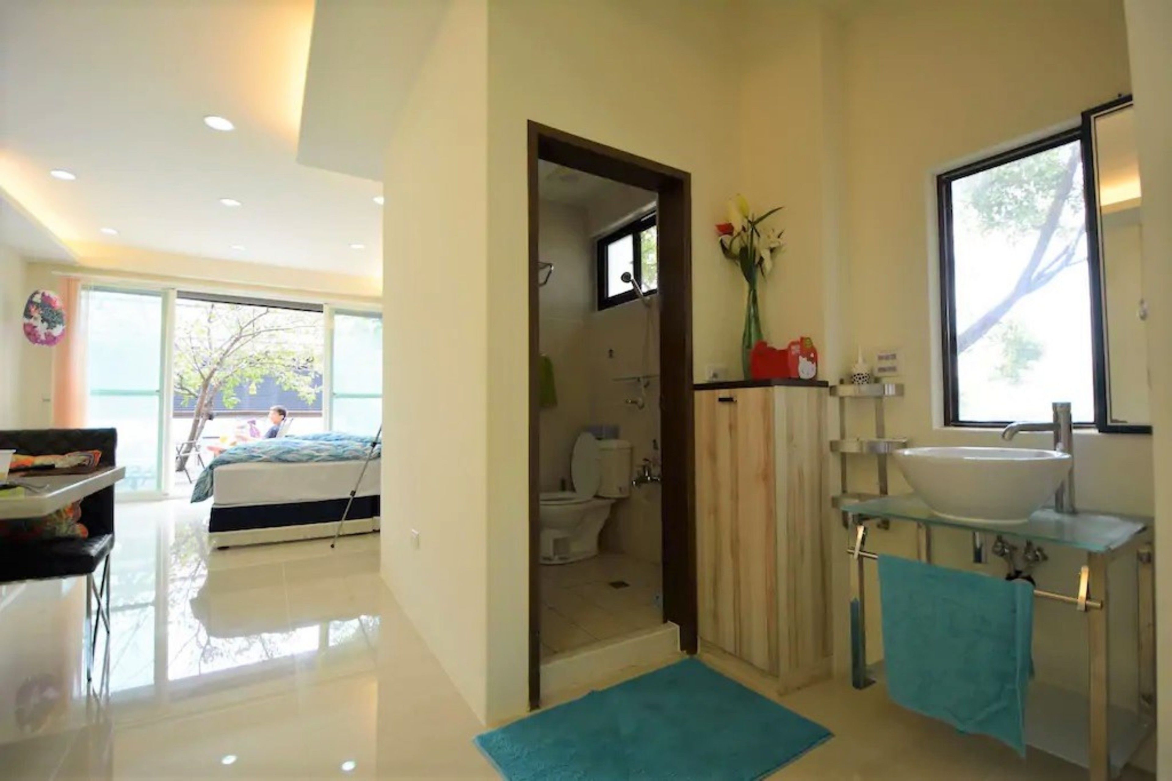 Un baño moderno con lavamanos de vidrio, ducha y retrete. Hay una toalla azul en el suelo y un florero con flores sobre el lavamanos.