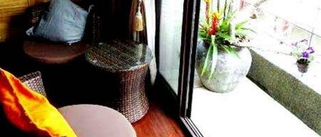 Una habitación con piso de madera, una silla de mimbre, una mesita y un ventanal con vistas al exterior.