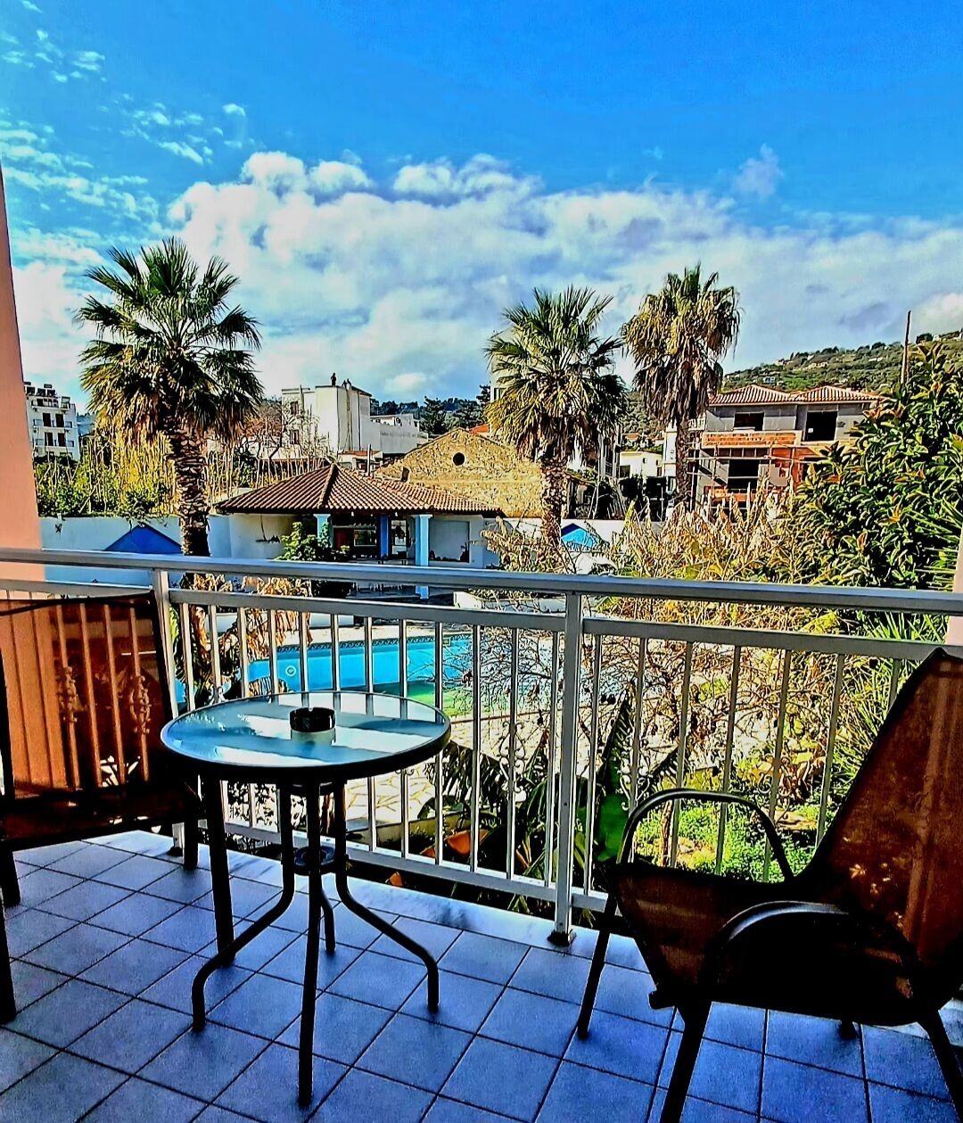 A balcony with a table and chairs, overlooking a swimming pool and a residential area with palm trees and houses.