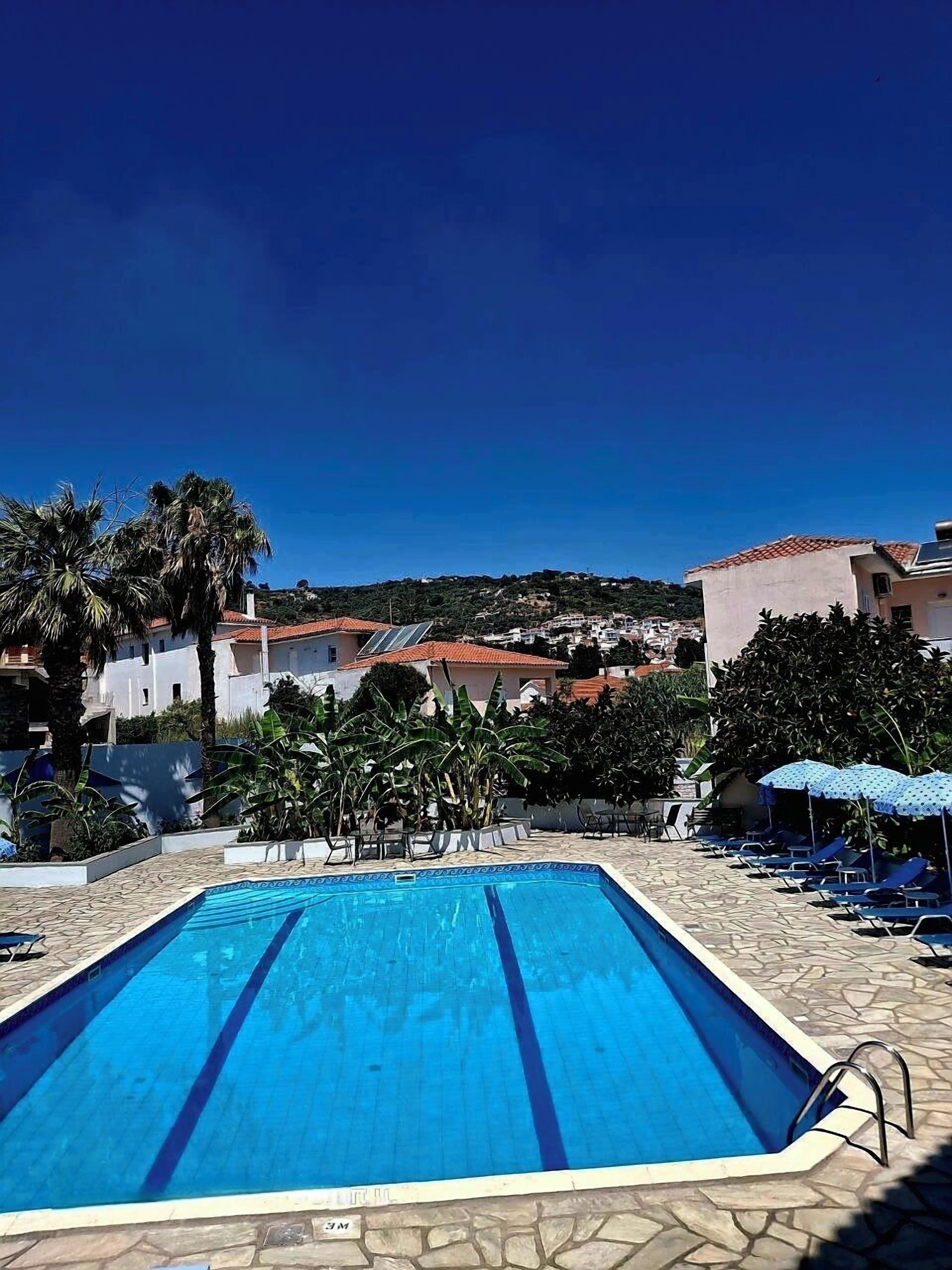 Una piscina con acqua cristallina e azzurra, circondata da lettini e ombrelloni, con edifici e alberi sullo sfondo.