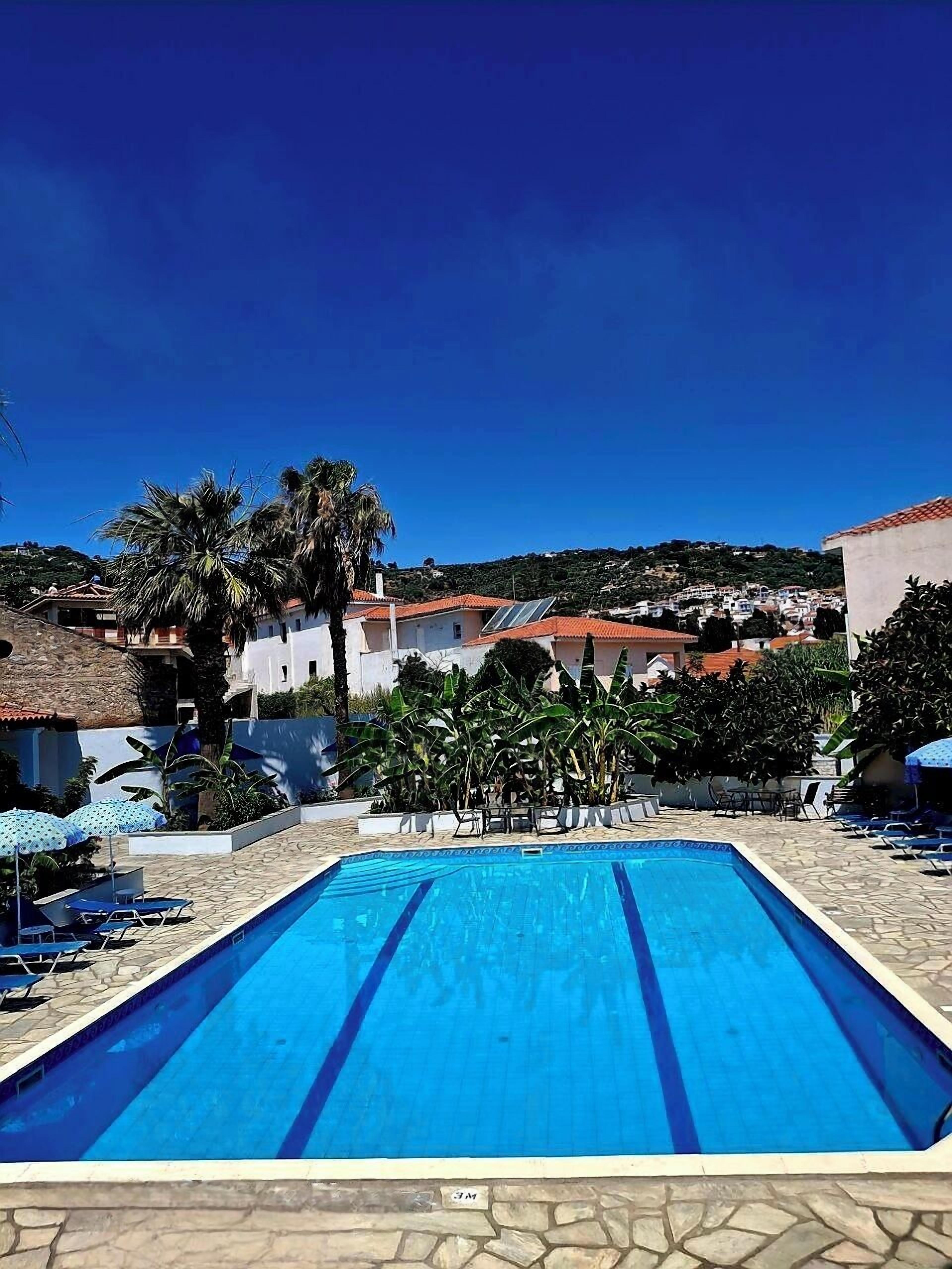 A swimming pool with lounge chairs and umbrellas, surrounded by palm trees and buildings.