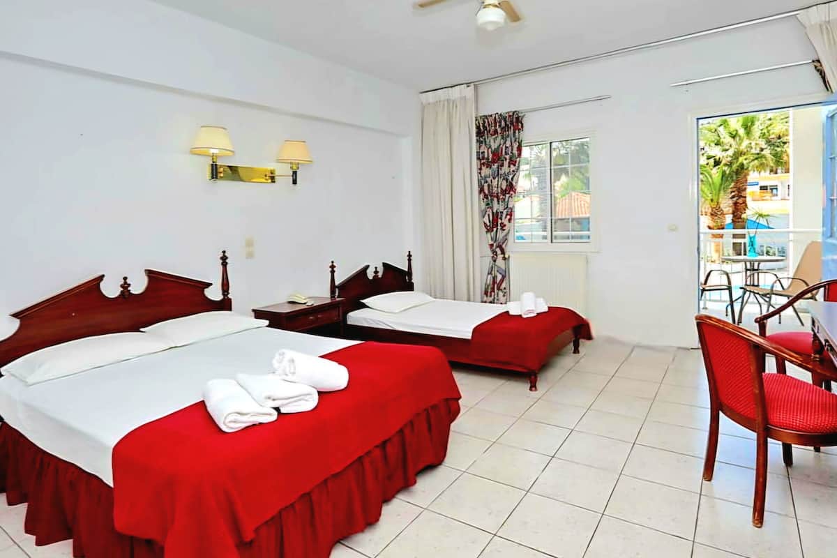 A hotel room with two beds, a ceiling fan, a window with curtains, a small table with a chair, and a view of the outside.