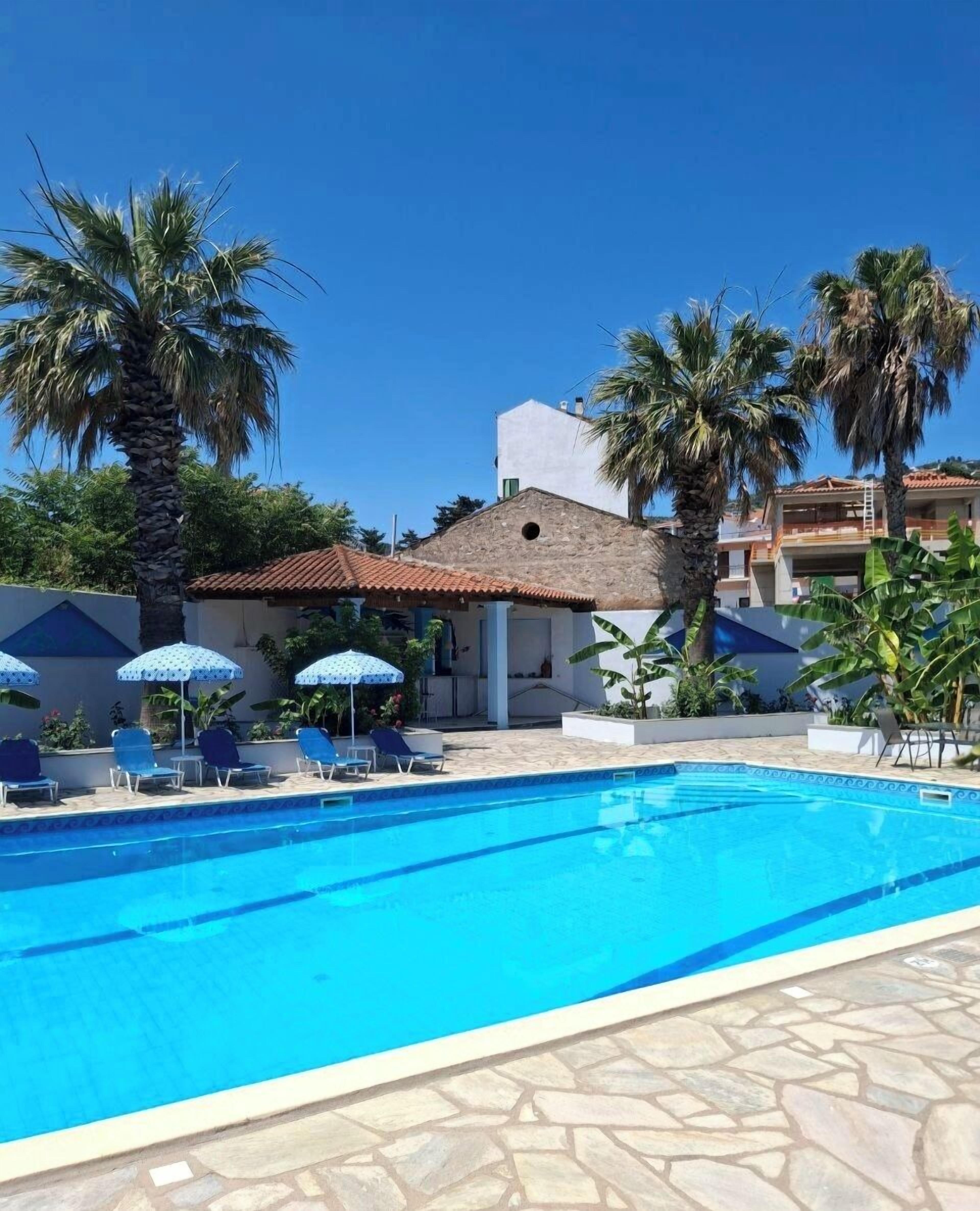 A swimming pool with lounge chairs and umbrellas, surrounded by palm trees and a building with a tiled roof.