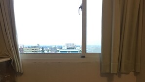 Ein kleines Zimmer mit Fenster und Blick auf die Stadtlandschaft, ein Holzstuhl, ein kleiner Tisch und eine Küchenzeile.