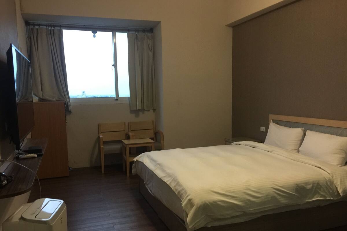 Una habitación de hotel con una cama, un escritorio con silla, un televisor y un ventanal con cortinas.