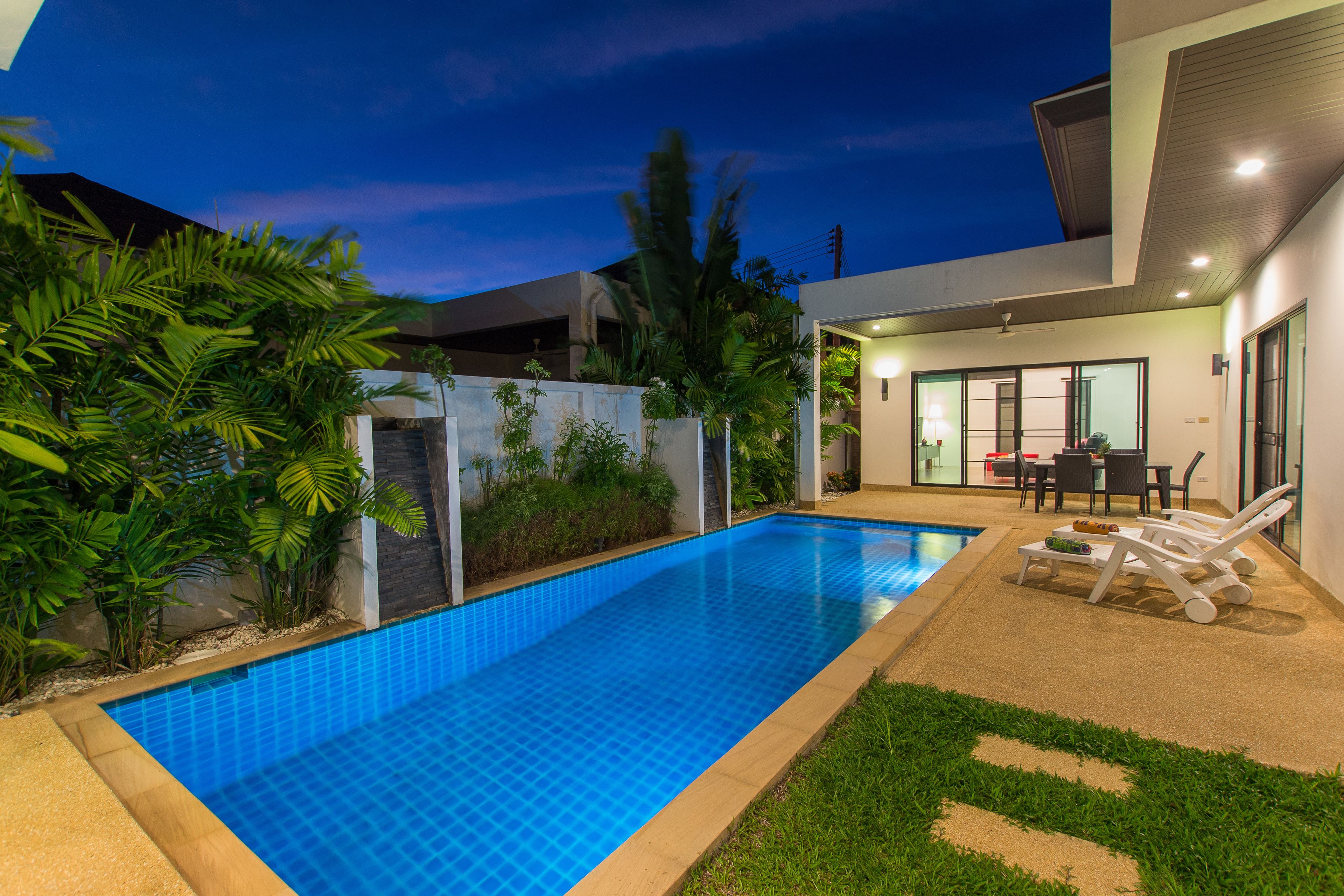 A modern outdoor pool area with a clear blue pool, surrounded by lush greenery and a well-maintained lawn.