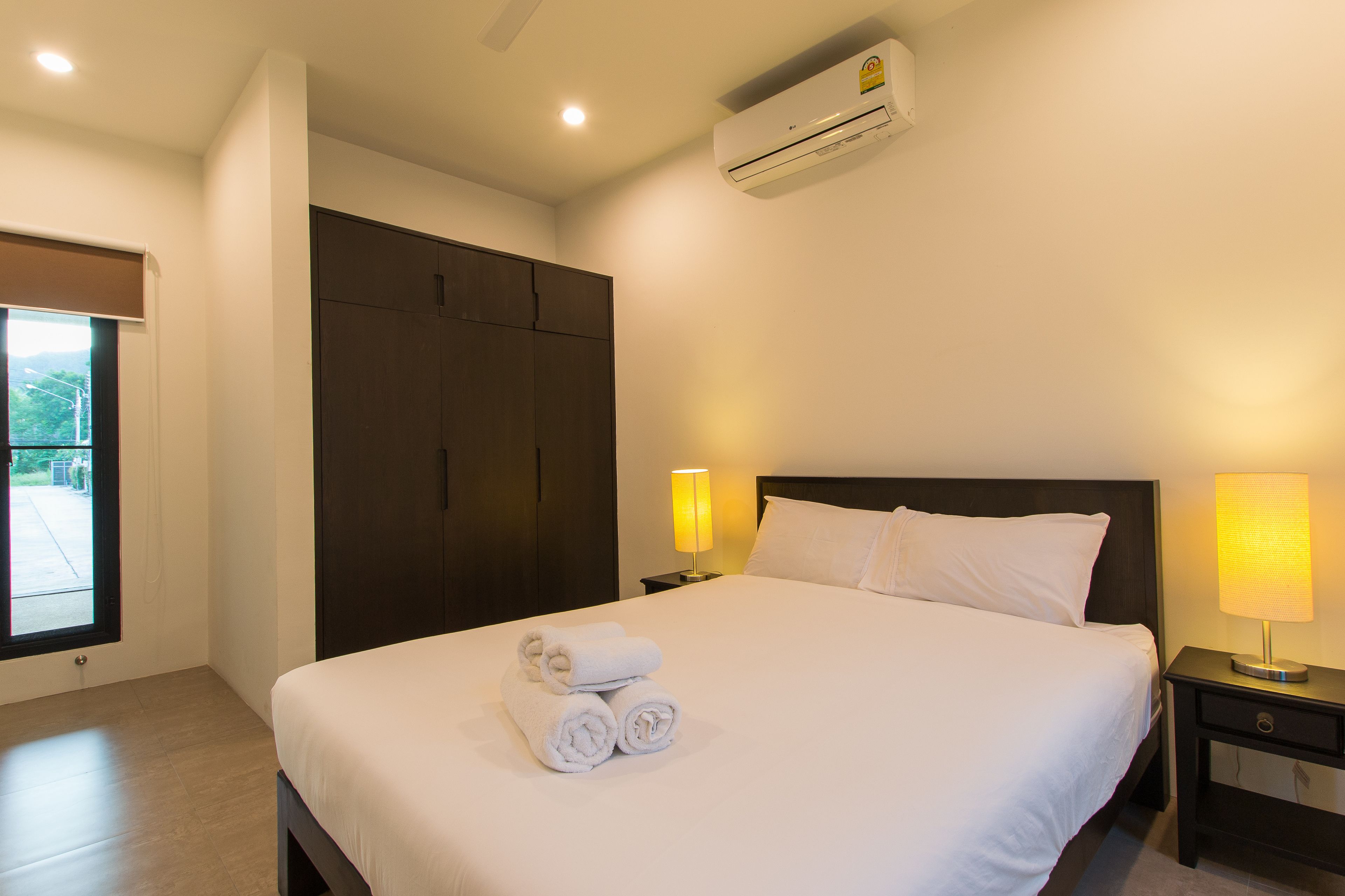 A hotel room with a large bed, a wardrobe, an air conditioning unit, a nightstand with a lamp, and a window with a view.