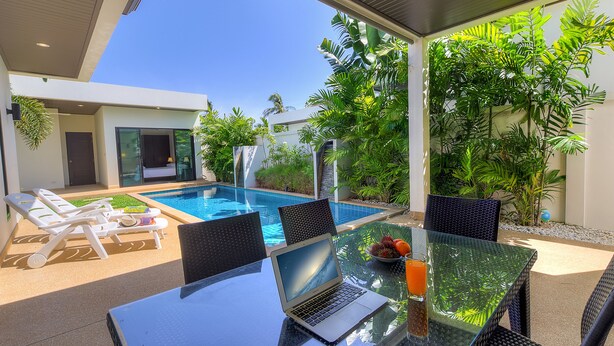 Een moderne buitenruimte met een glazen tafel, laptop en lounge fauteuils bij een zwembad.