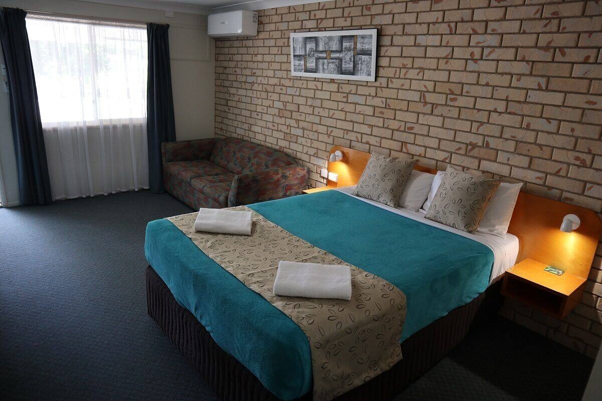 A hotel room with a bed, a sofa, a window with curtains, a brick wall, and a painting.