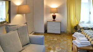 A hotel room with a sofa, a bedside table with a lamp, a bed with a patterned bedspread, and a window with yellow curtains. - B&B La Locanda dell'Arte Museo (Naples)