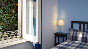 A bedroom with a bed, a nightstand, a lamp, a window with a view of greenery, and a door. - B&B La Locanda dell'Arte Museo (Naples)