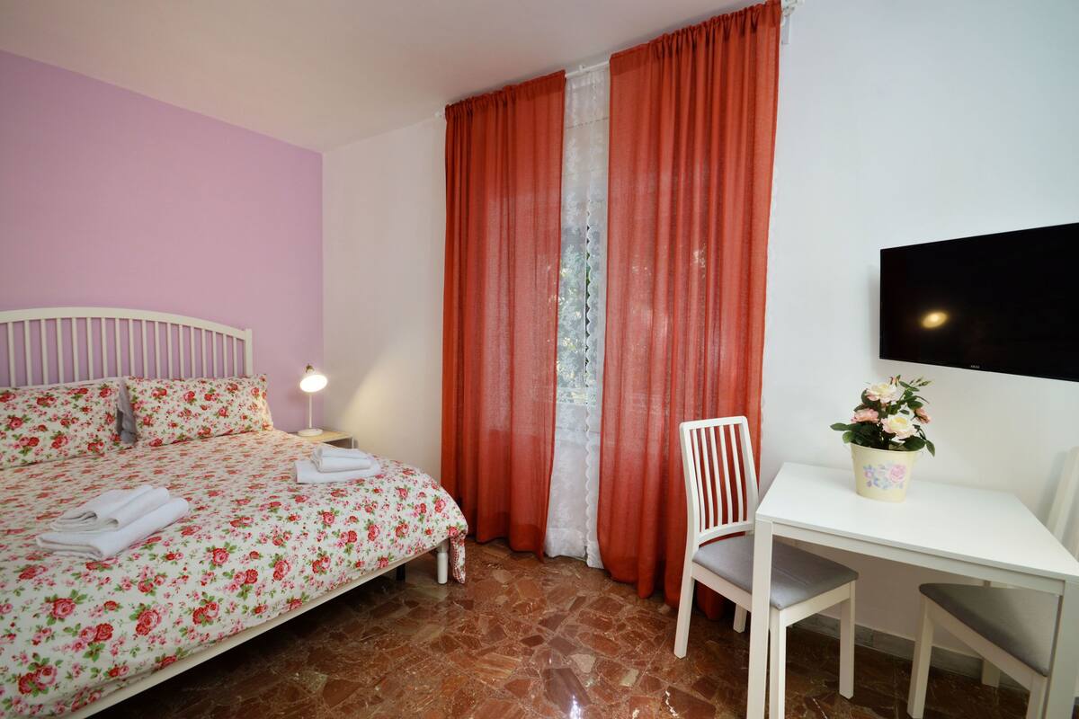A bedroom with a bed, a nightstand, a chair, a table, and a TV.
