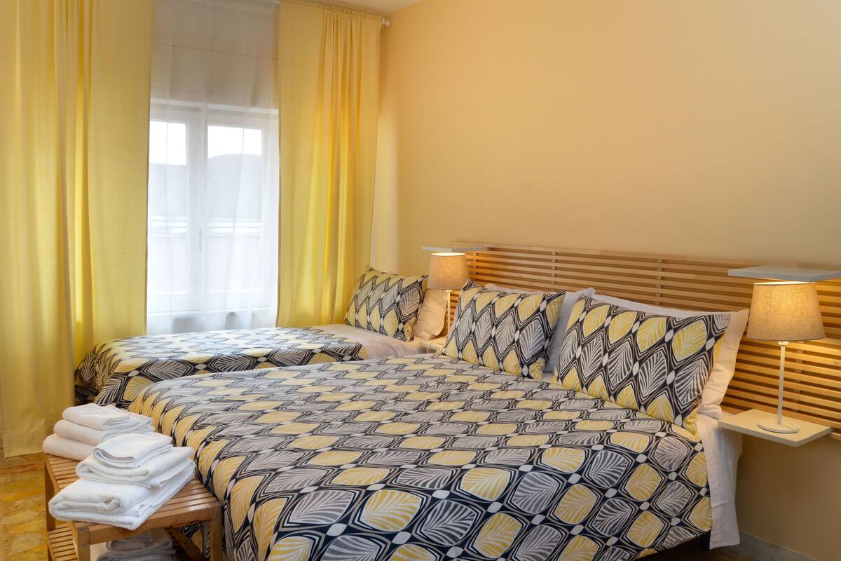 A double bed with a patterned bedspread, two bedside lamps, a wooden bench with towels, and a window with yellow curtains.