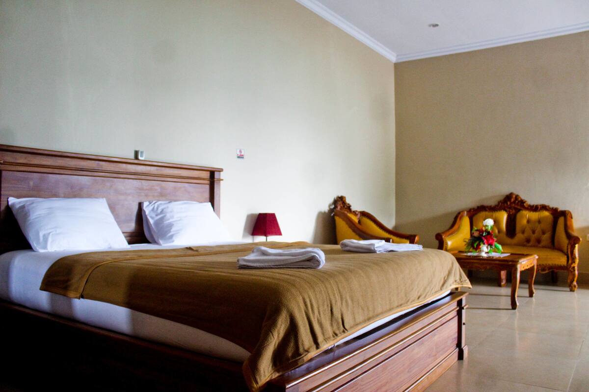 A hotel room with a large bed, a wooden headboard, a red lamp, a sofa, and a small table with a vase.