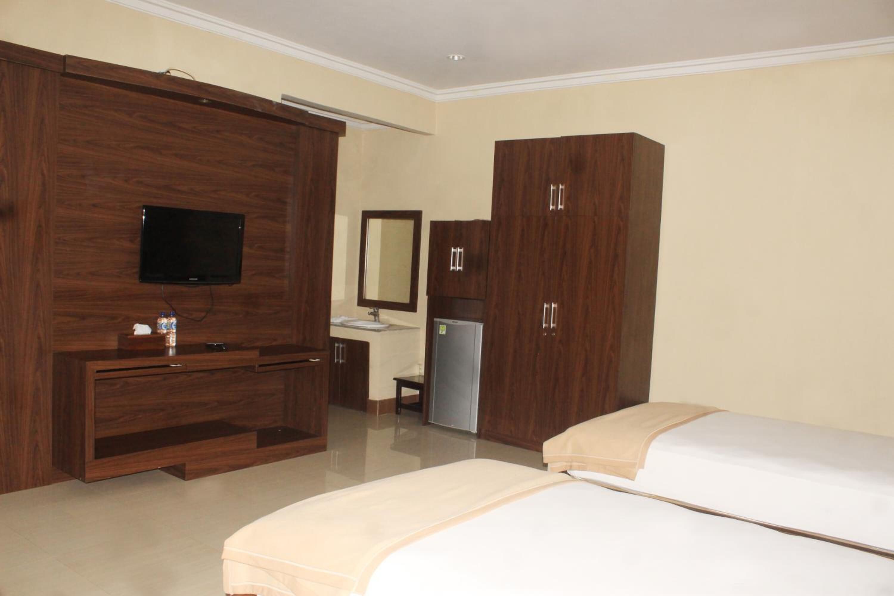 A hotel room with a large wooden wardrobe, a television mounted on the wall, a small desk with a mirror, and two beds.