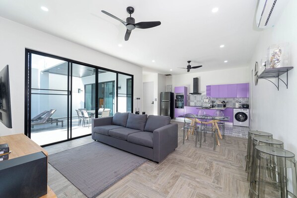 Un salon moderne avec un canapé gris, une table basse et un téléviseur. On y trouve également un coin cuisine avec des meubles violets et un coin repas avec une table et des chaises.