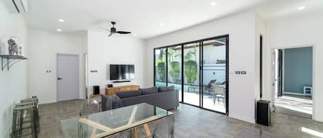 A spacious living area with a glass dining table, a gray sofa, a TV, and a ceiling fan.