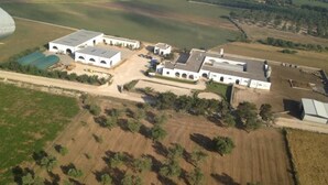 An aerial view of a large estate with multiple buildings, including a main house with a large roof and several smaller structures. There are open fields and scattered trees around the property. - Masseria Autigne (Otranto)