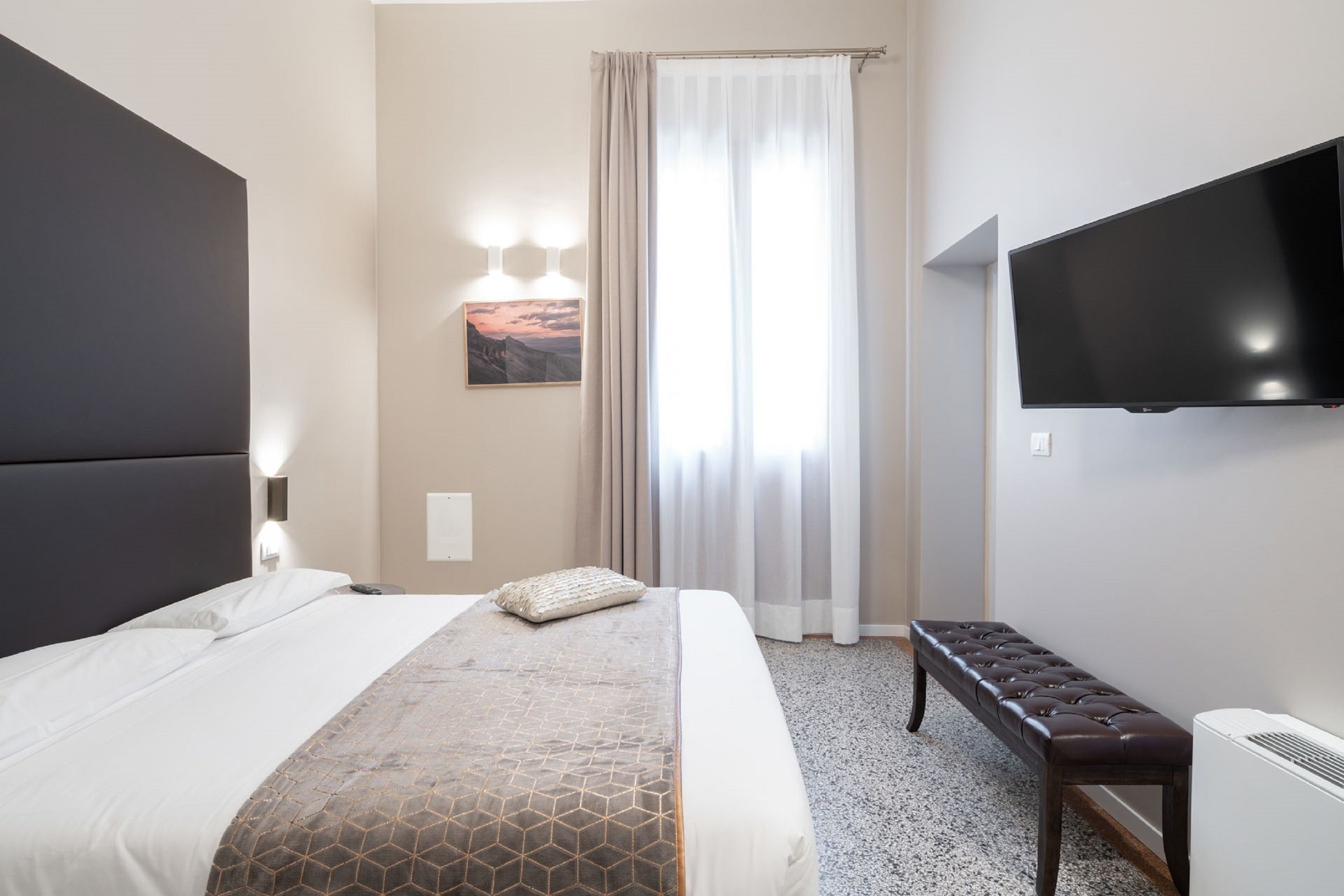 Una habitación de hotel con una cama grande, un banco, un televisor de pantalla plana y una ventana con cortinas.