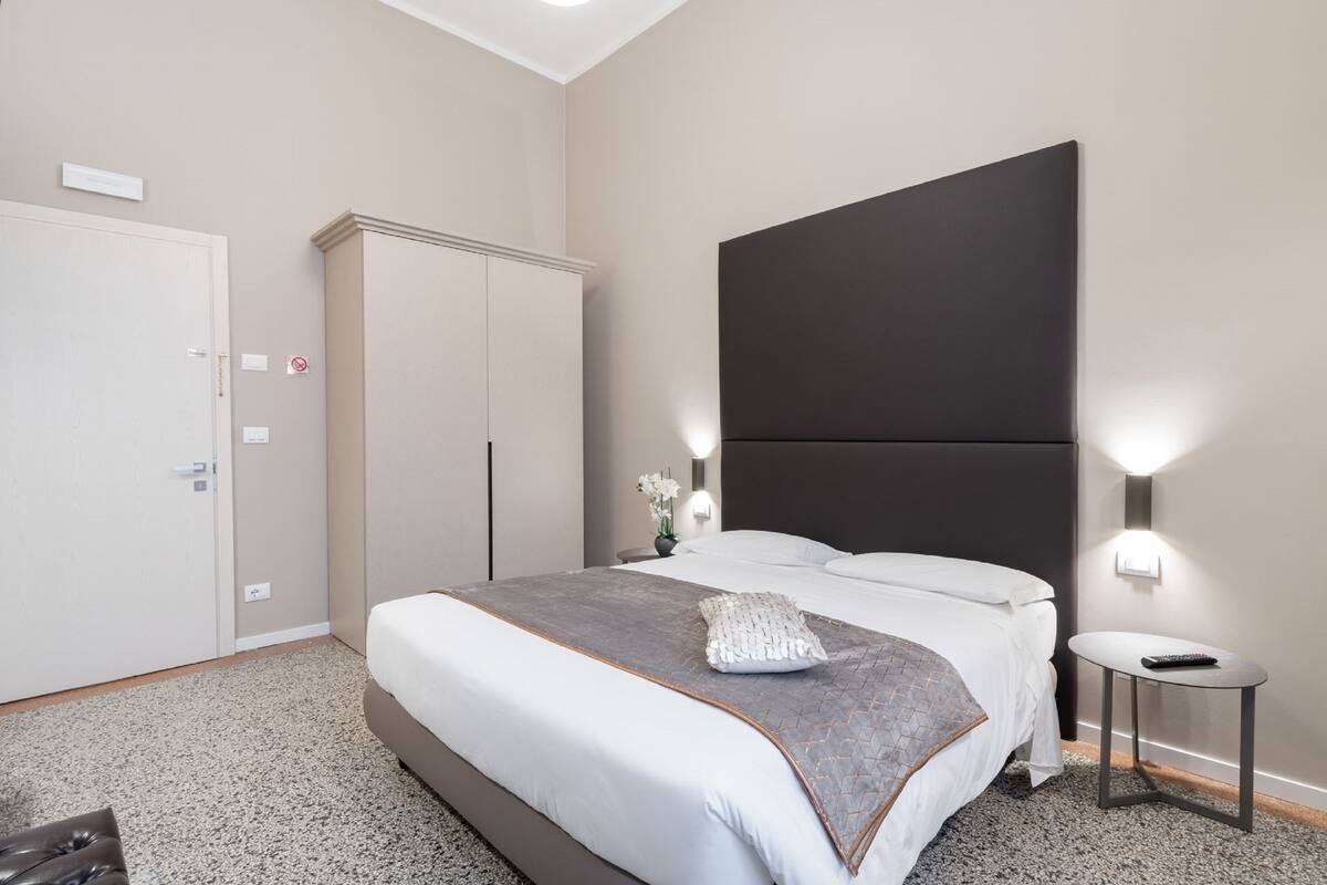 A modern hotel room with a large bed, a wardrobe, a small table, and a nightstand.