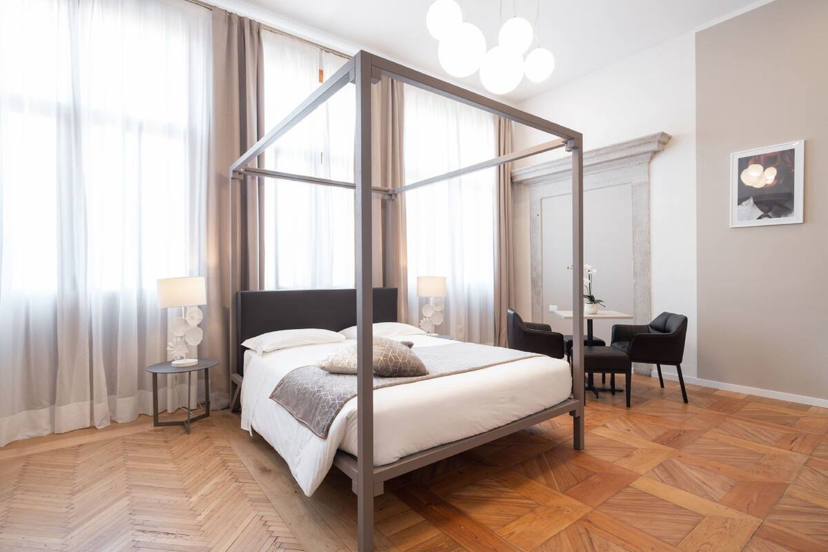 A modern hotel room with a four-poster bed, a wooden herringbone floor, a desk with a chair, and a large mirror on the wall.