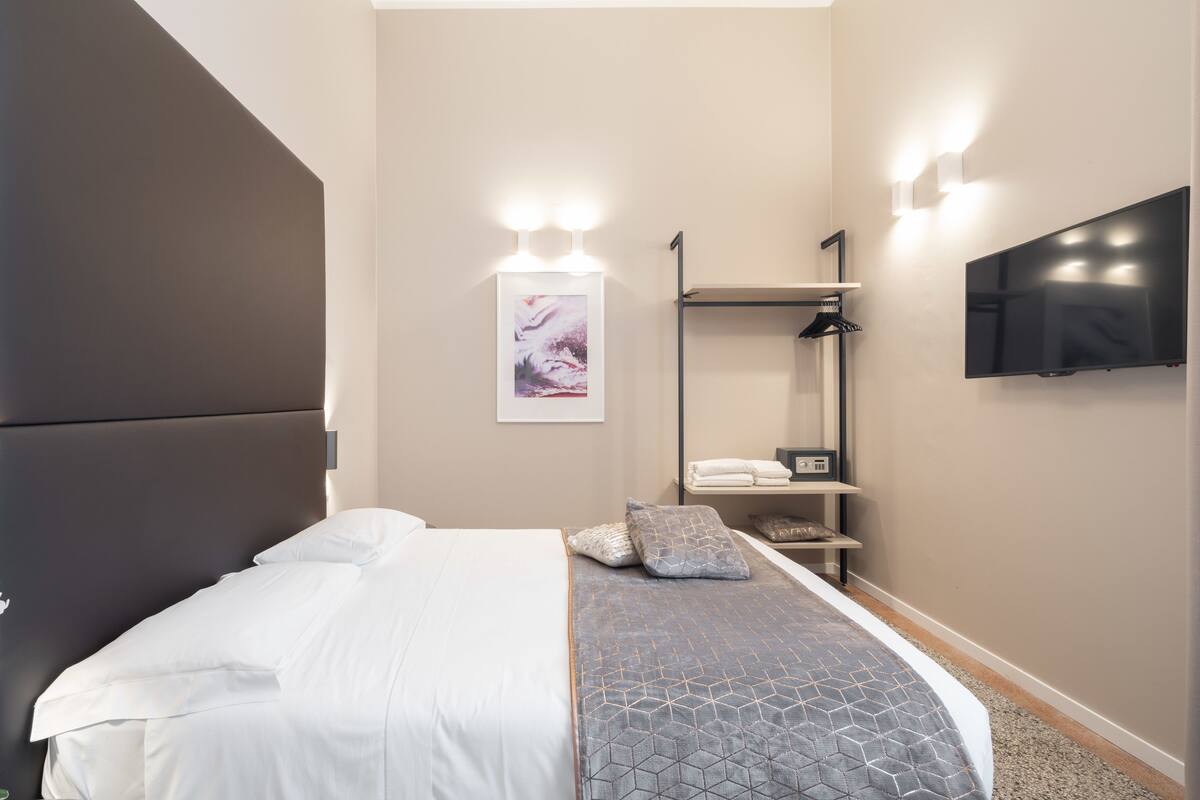 A modern hotel room with a bed, a flat-screen TV mounted on the wall, and a shelf with towels.