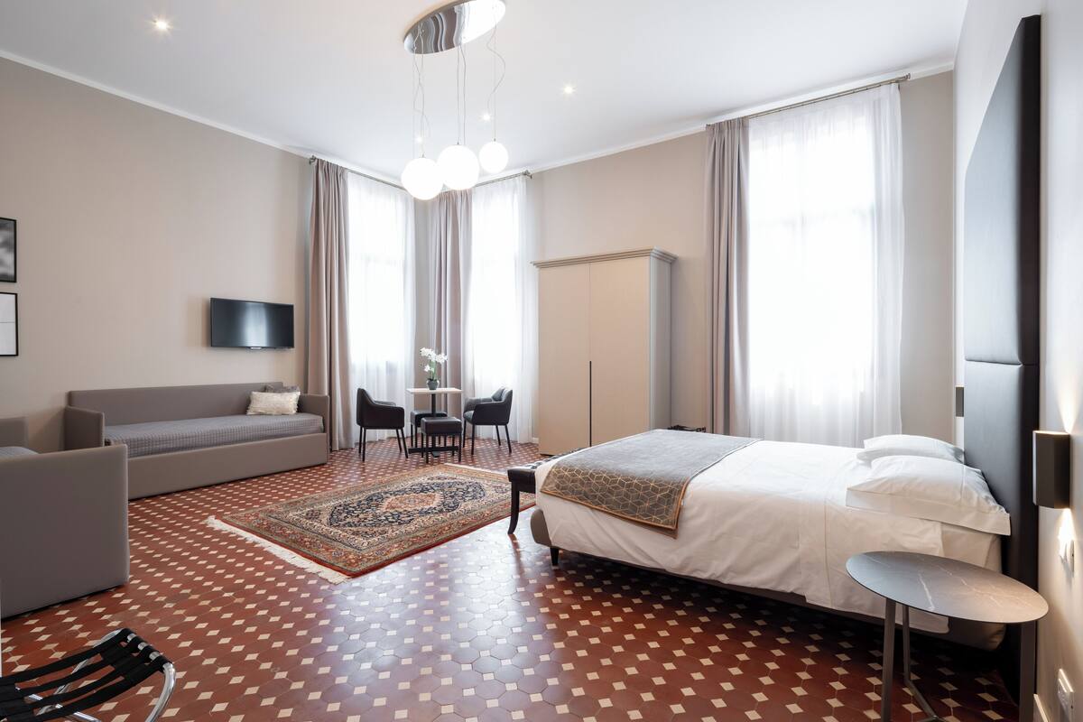 A modern hotel room with a large bed, a sofa, a TV, and a dining area.