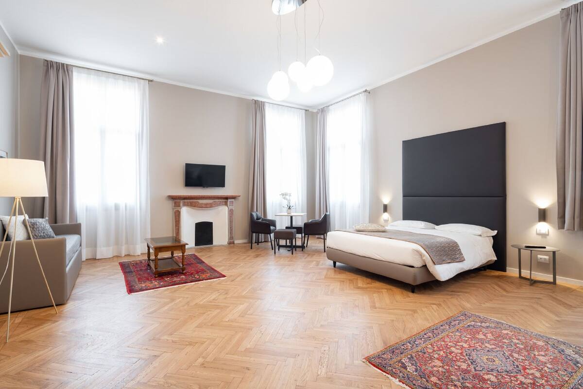 A modern hotel room with a large bed, a TV, a fireplace, a dining table with chairs, and a sofa.