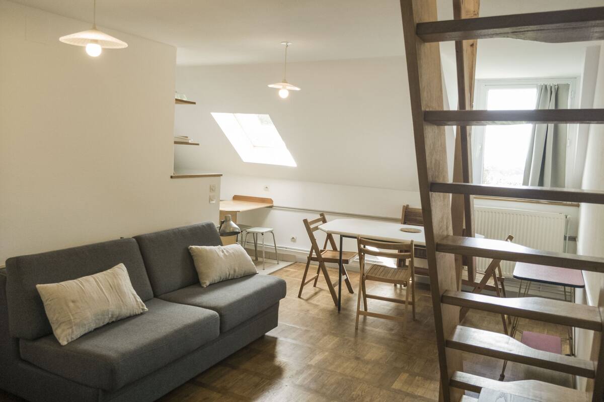 Una sala moderna con un sofá gris, una mesa de comedor de madera y sillas. Hay un tragaluz sobre la zona del comedor y una escalera de madera al frente.