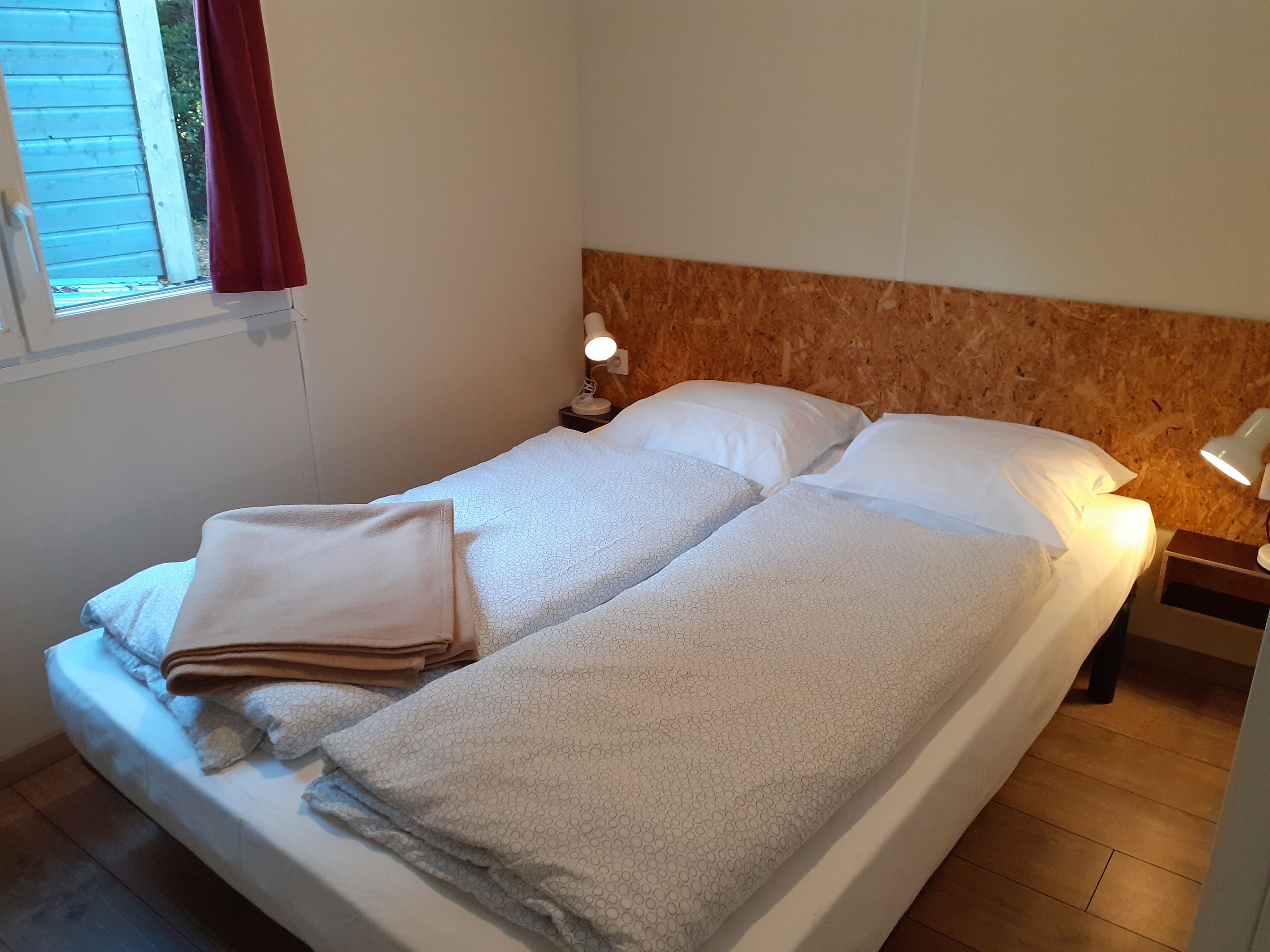 A single bed with a wooden headboard, two pillows, a bedside lamp, and a window with a red curtain.