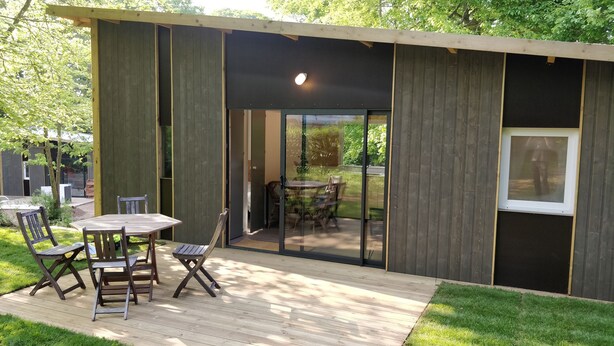Uma área moderna para refeições ao ar livre, com um deck de madeira, cadeiras de estrutura metálica e uma porta de vidro que dá para um jardim.