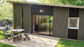 Uma área moderna para refeições ao ar livre, com um deck de madeira, cadeiras de estrutura metálica e uma porta de vidro que dá para um jardim.