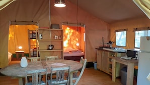 Uma tenda acolhedora com uma mesa de jantar de madeira, cadeiras, uma área de cozinha com fogão e uma cama com luz quente.