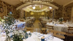 Uma sala de banquetes com mesas redondas preparadas para um evento formal, paredes de pedra e luzes de corda.