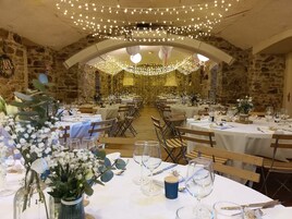Uma sala de banquetes com mesas redondas preparadas para um evento formal, paredes de pedra e luzes de corda.
