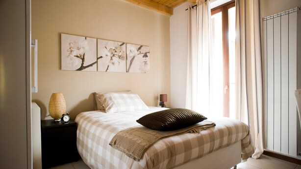 A bedroom with a bed, a nightstand with a lamp, a wall-mounted picture, and a window with curtains. - Camera 40 - In Gorgonzola (Gorgonzola)